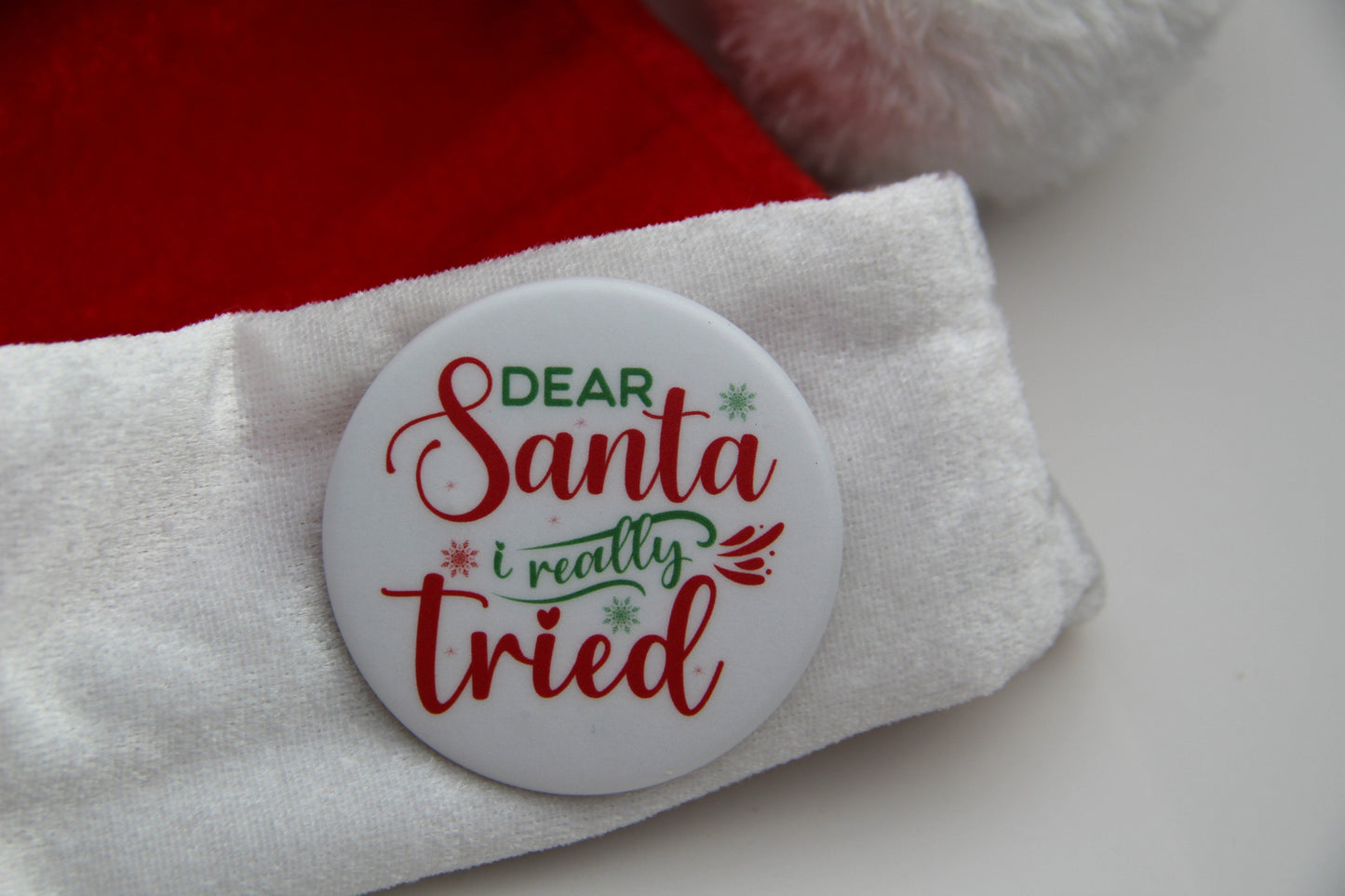 A white pinback button on a Santa hat with a saying. The button reads Dear Santa I really tried.