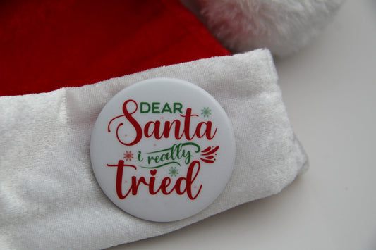 A white pinback button on a Santa hat with a saying. The button reads Dear Santa I really tried.